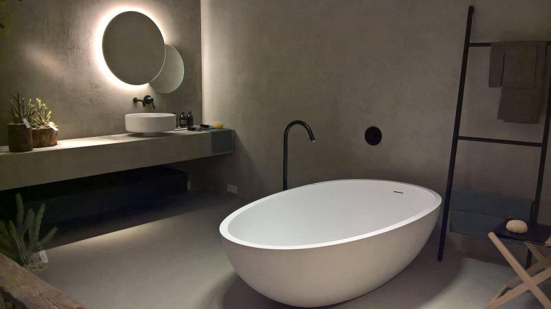 White oval bath on grey flooring with an illuminated mirror